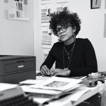 Lizania, a thirty-seven-year-old lighter skin Black woman with black short curly hair. With large glasses, large earrings sitting at her desk with her arms propped on it. You can see her typewriter as well as a black printer.